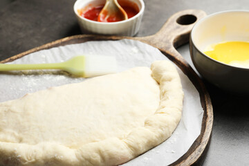 Uncooked calzone pizza, sauce and egg yolk on grey table, closeup
