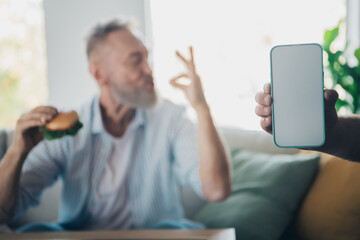 Elderly man enjoying a burger while casually relaxing indoors on a bright day, showcasing comfortable leisure in a home setting.