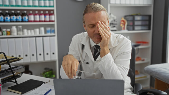Mature caucasian man in a clinic room appears stressed with hand on face, surrounded by medical files and bottles, suggesting a busy healthcare professional environment.