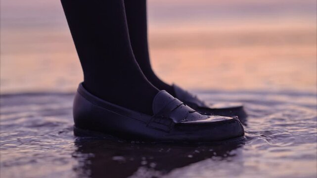 朝焼けの海岸で波打ち際に立つ女子高校生の足元