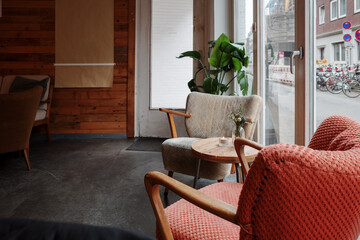 A warm and inviting café interior featuring vintage armchairs with textured upholstery, a wooden table with a small flower arrangement, and large windows allowing natural light to enter.