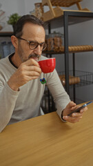 Mature man in an office setting holding a red cup while using a smartphone, showcasing a blend of work relaxation within an organized and modern workplace interior.