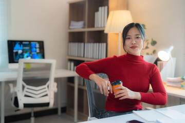 Asian businesswoman in red turtleneck sweater holding coffee cup while sitting at desk in modern office enjoying break time, computer with graphs and charts on screen in background