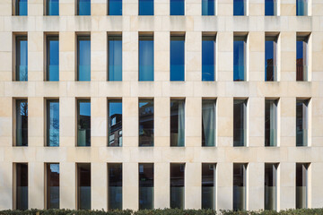 A contemporary building exterior featuring a beige stone facade with a repetitive pattern of narrow, rectangular windows.