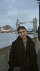 Woman smiling on london street with iconic bridge in the background, wearing stylish winter clothing, capturing vibrant city atmosphere during a cloudy day.