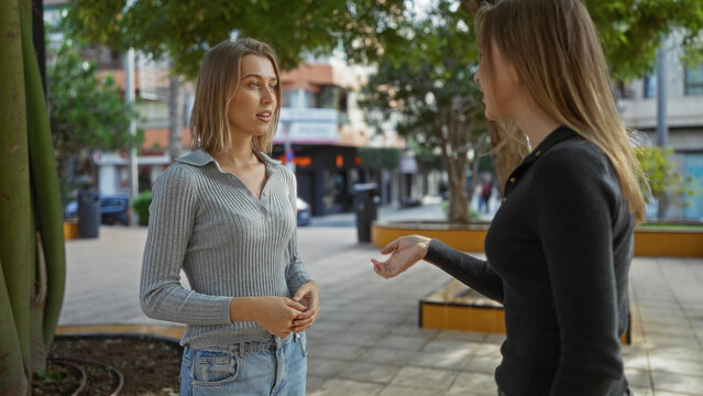 Women talking in an urban park setting, showcasing family bond and interaction, with trees and city street in background, reflecting sisterly love and connection outdoors.