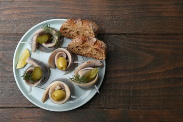 Delicious herring rolls with olives, pickles, dill and bread pieces on wooden table, top view....