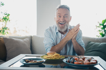 Cheerful elderly man enjoying a weekend meal at home, featuring a variety of food dishes and a comfortable living room setting