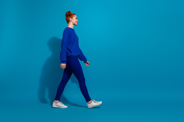 Young woman walking in front of blue background wearing fashionable outfit featuring casual blue knit sweater and pants.