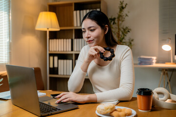 Asian businesswoman eating donuts and using laptop computer while working late at night in home office