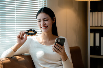 Happy asian woman enjoying her break time with donut and mobile phone