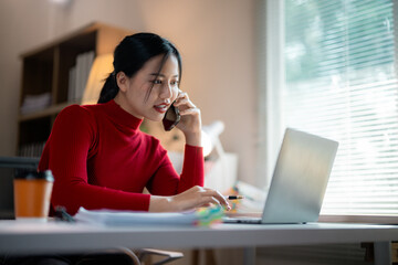 Young Asian businesswoman wearing red turtleneck sweater talking on mobile phone and working on laptop in home office