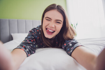 Naklejka premium Young woman winking playfully while lying on bed in cozy pajamas, expressing cheerful emotions and relaxation