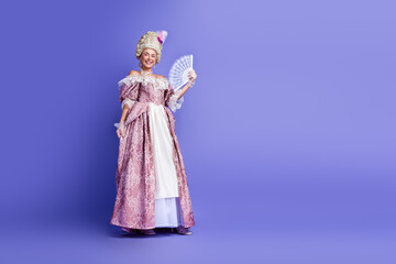 Fototapeta premium Elegant woman in historical baroque costume posing with a fan against a vivid violet studio background