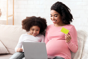 Online Shopping Concept. Pregnant Afro Mom With Laptop And Credit Card Purchasing Baby Clothes In...