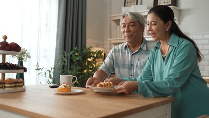 Energetic grandmother holding breakfast and serving to her husband while grandfather sitting at modern kitchen. Smiling elder couple spending time together. Retirement lifestyle concept. Myrmidon.