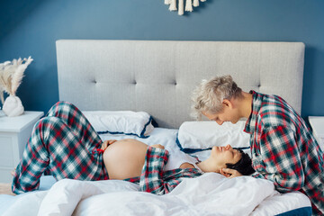Mixed race couple are waiting for a baby. Husband and his pregnant wife in cozy pajamas relaxing on the bed at modern home bedroom. Romantic moments for pregnant couple, warm atmosphere of love.