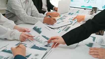 A business meeting scene showing a diverse team engaging with data charts and reports on the table, alongside laptops and digital screens in a modern office setting. SACTR