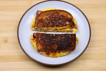 A homemade breakfast sandwich cut in half with crispy toasted bread, scrambled eggs, bacon, and melted cheese on a plate over a wooden surface. A rustic, hearty meal perfect for breakfast or brunch.