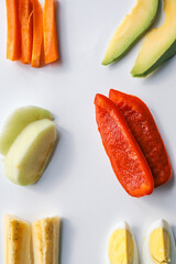 Assorted fresh food ingredients arranged on a white background, including carrot sticks, avocado slices, apple pieces, red bell pepper, banana chunks, and boiled egg halves