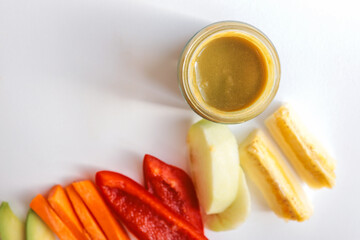 A glass jar of homemade baby puree with fresh vegetables and fruits arranged on a white background. Includes carrot, bell pepper, apple, and banana. Healthy organic food for infants and toddlers