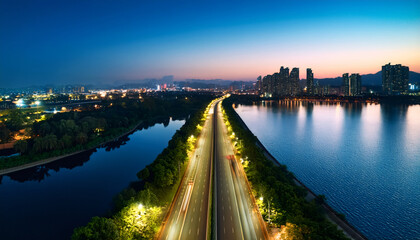 Obraz premium Cityscape at Dusk: An elevated perspective captures a city skyline at dusk. a serene expanse of water surrounds a pathway with illuminated light trails and a vibrant sky.