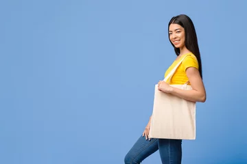 Joyful Young Asian Woman With Blank Textile Tote Eco Canvas Shopping Bag Posing Over Purple Background In Studio, Smiling At Camera, Empty Copy Space For Your Logo, Advertisement Or Branding © Prostock-studio