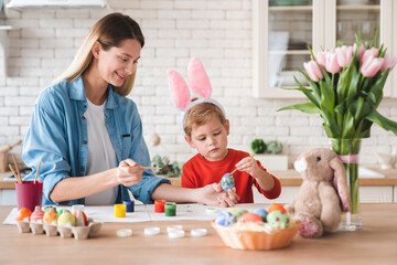 Fototapeta premium Happy positive mother and little son decorating eggs for Easter at home. Mom and her small kid child painting ornaments for holiday celebration together in the kitchen