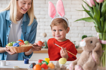 Nice little boy in bunny ears for Easter painting eggs in the kitchen with his mom. Small kid child...
