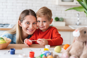 Naklejka premium Cheerful caucasian mother and her little son painting eggs for Easter at home. Portrait of happy mom and kid child boy looking at camera while decorating for spring festive celebration