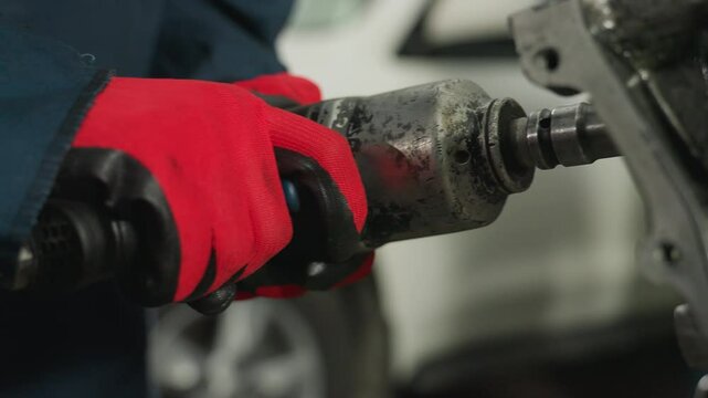 Mechanic wearing red gloves operates pneumatic tool on engine block in industrial workshop, close-up captures precision repair work and metal components with blurred white vehicle in background