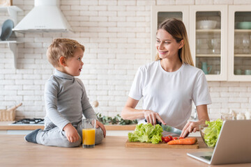 Little small kid child son helping his mom in cooking healthy fresh food meal in the kitchen drinking orange juice. Loving mother preparing vegetable salad with her son