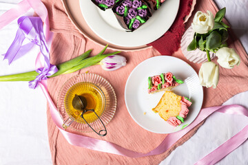 Muffins with candied fruits and cream in the form of tulips on a white plate, homemade pastries for Mother's Day, light composition, muffins with tea, tulips and pink silk ribbons in the background