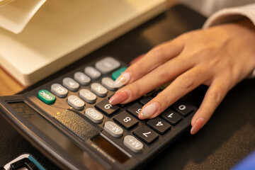 A woman is sitting at a table with a package in front of her. She is calculating expenses in a notebook and laptop and talking on her mobile phone.

