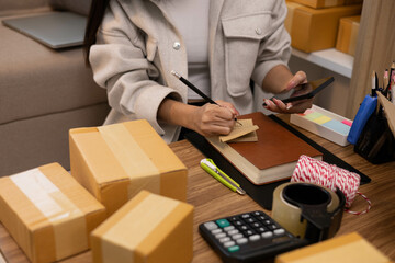 A woman is sitting at a table with a package in front of her. She is calculating expenses in a notebook and laptop and talking on her mobile phone.
