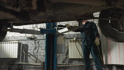 Mechanic in dark uniform and gloves operating hydraulic lift in mechanical workshop, raising a car for inspection, underneath view shows vehicle suspension, tires, and metal structure