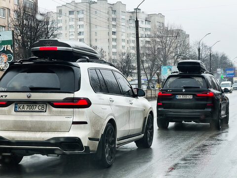 Two BMW X7 SUVs driving on a rainy urban road with rooftop cargo carriers