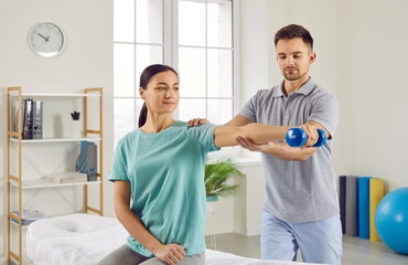 Obraz premium Young happy smiling woman patient doing sport exercise using dumbbells with support from male physiotherapist doctor in rehab clinic. Physiotherapy and physical rehabilitation concept.