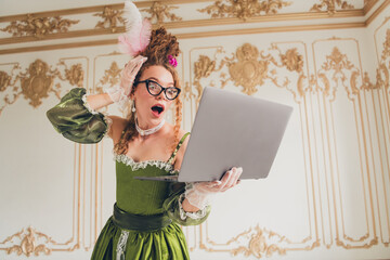 Elegant woman in a historic dress surprised while using a modern laptop indoors, merging past and...