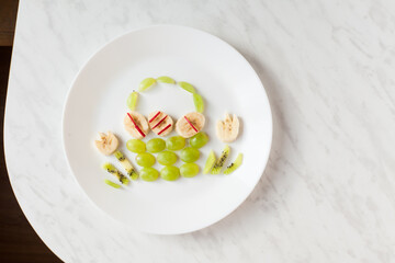 Creative healthy snack of breakfast for kids fruit bunny shaped on white plate, easter eggs around. Rabbit, basket makes from grape, orange, banana, apple. Top view