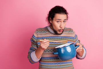 Young man with curly hair tasting soup from a pot in front of a vibrant pink background, showcasing...