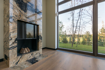 Fototapeta premium Fireplace decorated with marble tiles in the interior of the living room of a private house. Large panoramic window with a view of trees and a green lawn.