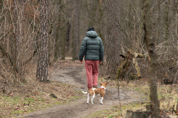 A person wearing a green jacket and red pants strolls down a dirt trail in a forest, accompanied by a small dog. The atmosphere is serene and tranquil, with bare trees around © Oleksandr Zinchenko