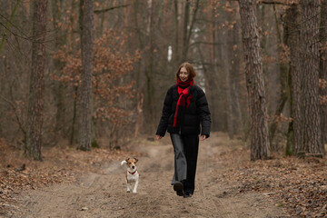 A woman in a black jacket and red scarf strolls down a dirt path in a forest, accompanied by her...