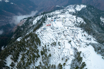A stunning aerial view of Shogran Hill Station blanketed in snow, with scattered buildings, winding roads, and dense evergreen forests on a mountain slope.
