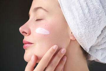 Side profile of a woman wearing a towel and gently applying moisturizing cream to her cheek with...
