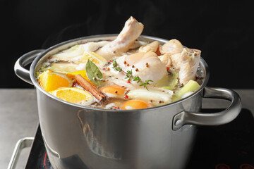 Whole fresh turkey, cut citrus fruits, thyme and spices boiling in pot on stove against black background, closeup