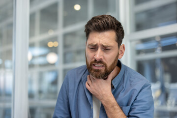Obraz premium A worried man dressed in blue casual shirt holds his neck expressing discomfort and illness, possibly suffering from a sore throat.