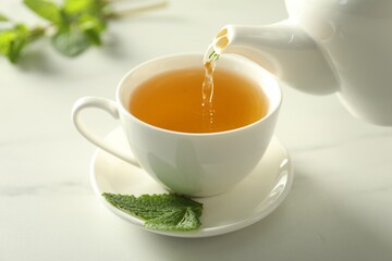 Pouring freshly brewed tea from teapot into cup at white marble table, closeup