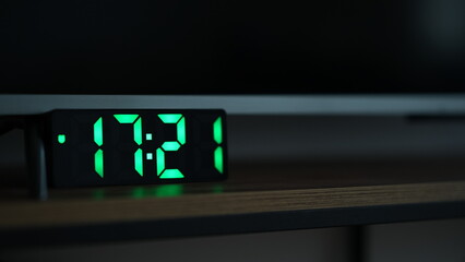 Green digital clock displaying time, placed next to a television. Modern LED alarm clock with bright numbers in a dimly lit room